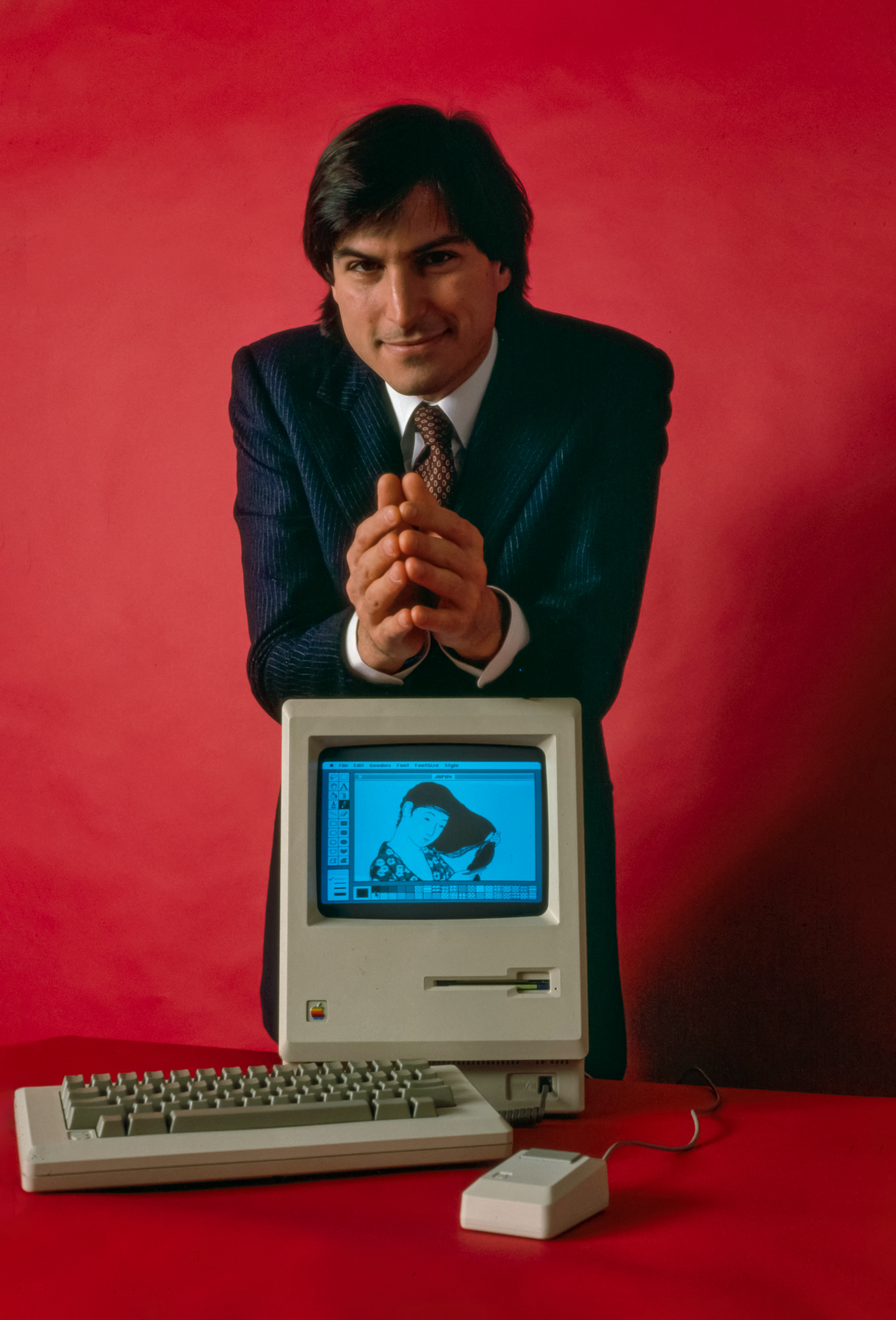 Steve Jobs posing in front of a Macintosh computer in 1984 on the screen is a drawing made with MacPaint of a woman combing her hair