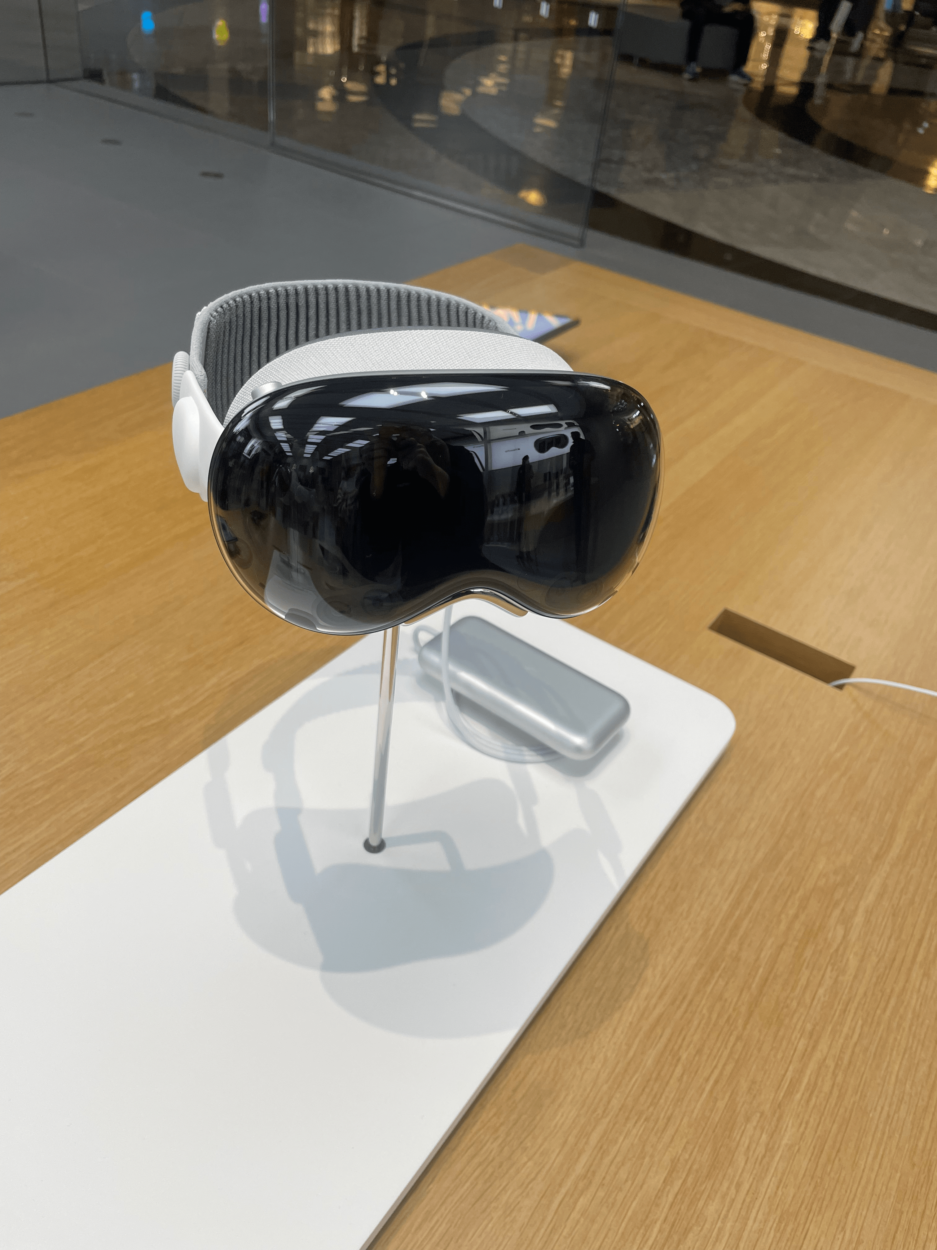 On a table at an Apple Store, the Apple Vision Pro VR headset, with black front glass and a white head strap, is pictured.