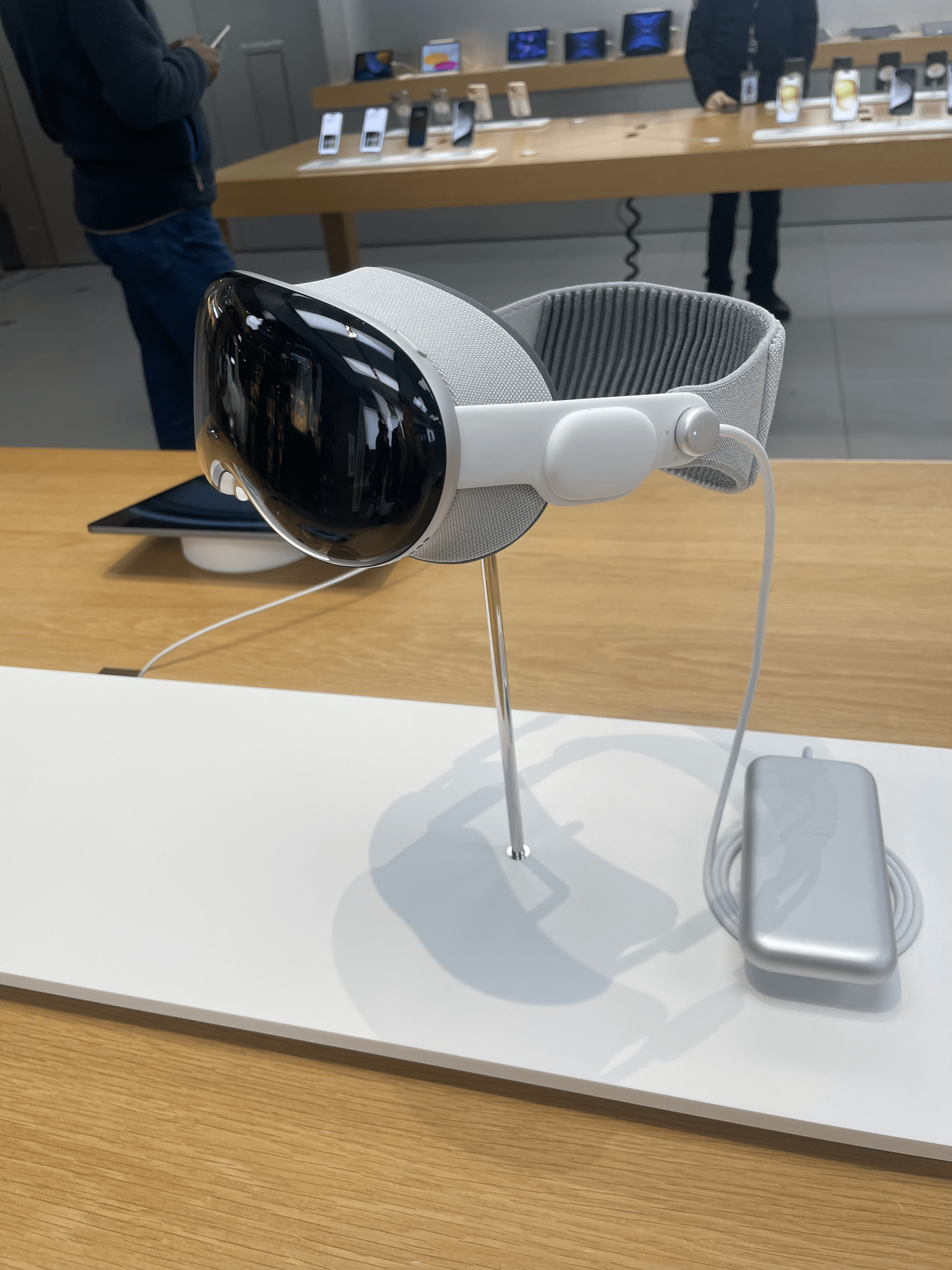 On a table at an Apple Store, the Apple Vision Pro VR headset, with black front glass and a white head strap with the light seal visible, is pictured.